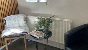 A room with a wooden slatted wall, a window with a plant, a radiator, a small table with a plant and a magazine, a fluffy chair, and a black sofa. - Hytten (Lemvig)