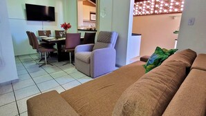 Ein Wohnzimmer mit braunem Sofa, einem Esstisch mit Stühlen, einem an der Wand befestigten Fernseher und einer durch einen offenen Durchgang sichtbaren Küche.