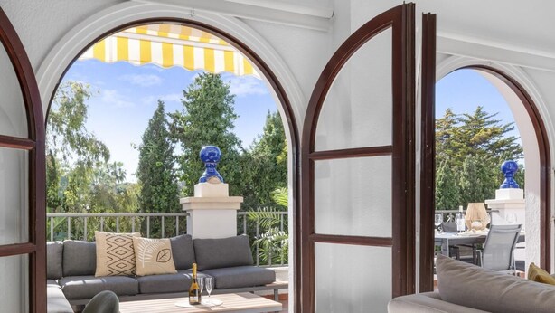 A sunlit patio with a sofa, a table, and a view of a garden and trees through arched openings. - Apartment in Andalucía del Mar for 4 people | ADM- 9-105 (Puerto Banús - Marbella)