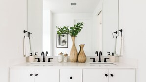 Une salle de bain moderne avec un meuble-lavabo blanc, deux robinets noirs et un grand miroir au-dessus du lavabo. On trouve deux vases en bois sur le meuble-lavabo et un tableau encadré au mur.
