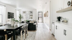 Une cuisine moderne avec des meubles blancs, un coin repas doté d’une table et de chaises noires, et un salon avec un canapé et des plantes.