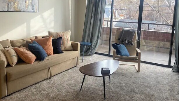 A living room with a beige sofa, a coffee table, and two armchairs. There is a large window with curtains, and a painting on the wall. - Charming 1 Bed Condo, Only 15 Minute Walk to Downtown (Salt Lake City)