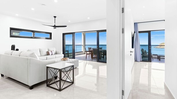 A modern living room with a white sofa, a coffee table, and a ceiling fan. There is a large window with a view of the sea. - Indigo Oceanfront Condo Middle Floor - Unit 2, Indigo Oceanview Terrace 2 (Sint Maarten)