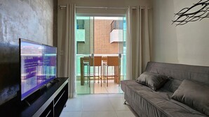 A living room with a flat-screen TV, a sofa, and a sliding glass door leading to a balcony with wicker furniture. - Lindo Apartamento na Praia do Forte! (204) (Cabo Frio)