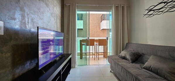 A living room with a flat-screen TV, a sofa, and a sliding glass door leading to a balcony with wicker furniture. - Lindo Apartamento na Praia do Forte! (204) (Cabo Frio)