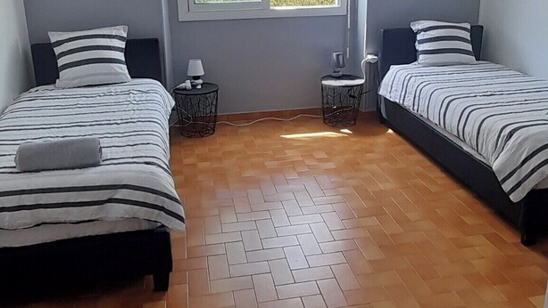 A room with two beds, a window, a ceiling light, and a herringbone-patterned floor.
