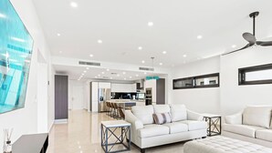 Een moderne woonkamer met een witte bank, een passende voetenbankje en een eethoek, met een keuken op de achtergrond.