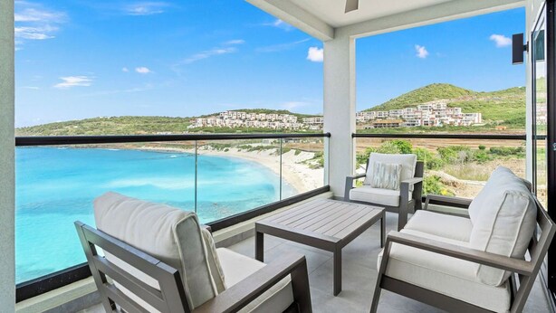 Een balkon met uitzicht op een strand en een heuvel. Er staan twee stoelen en een tafel.