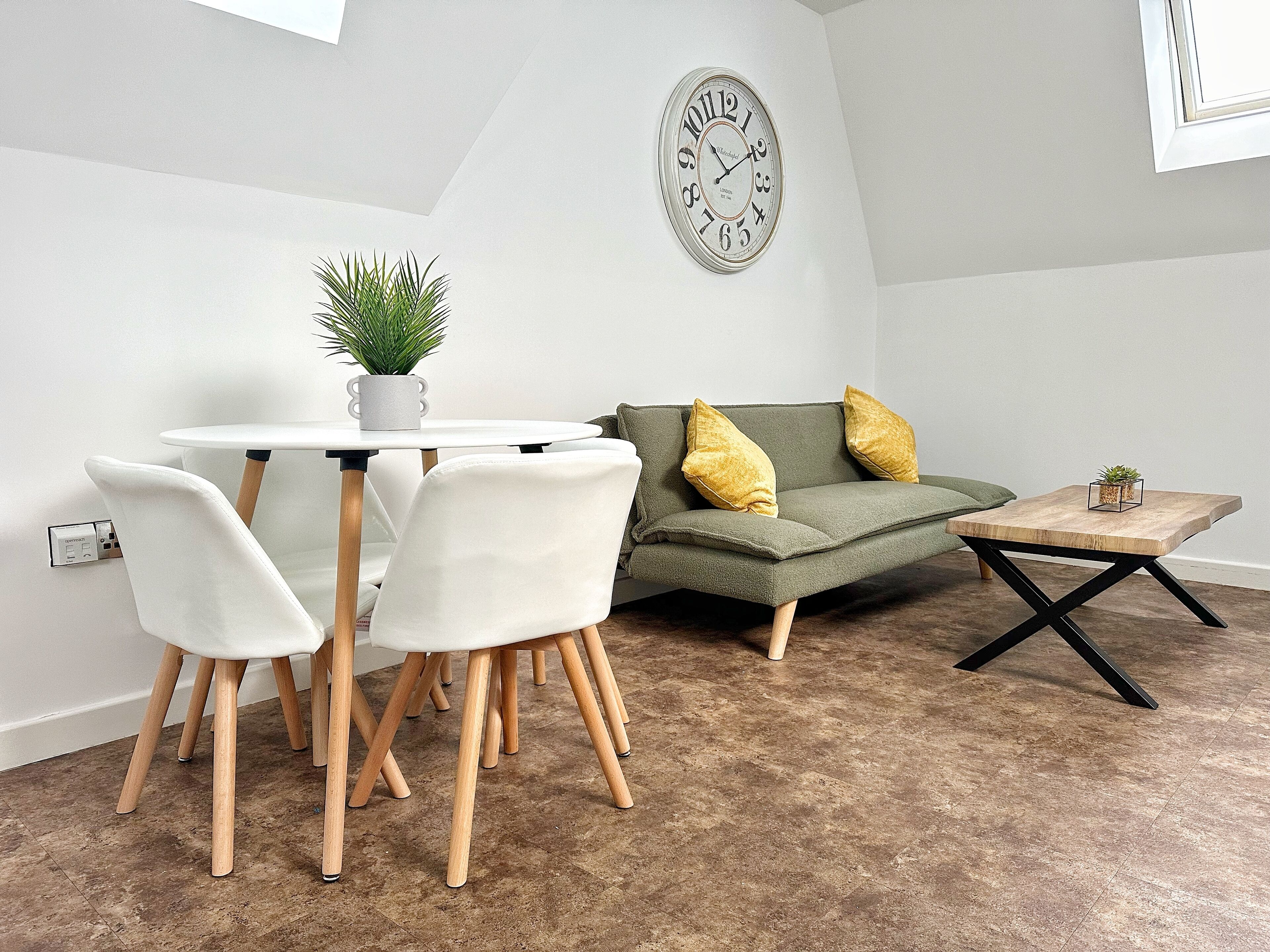 Ein modernes Wohnzimmer mit einem runden Esstisch, weißen Stühlen, einem grünen Sofa mit gelben Kissen und einem Holztisch.