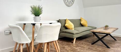 Ein modernes Wohnzimmer mit einem runden Esstisch, weißen Stühlen, einem grünen Sofa mit gelben Kissen und einem Holztisch.