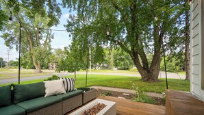 A wooden deck with a green sofa and a planter, surrounded by trees and a street view. - Charming 3-bedroom house with AC and WiFi. LAKE ACCESS King Beds! Newly Remodel (Minneapolis)