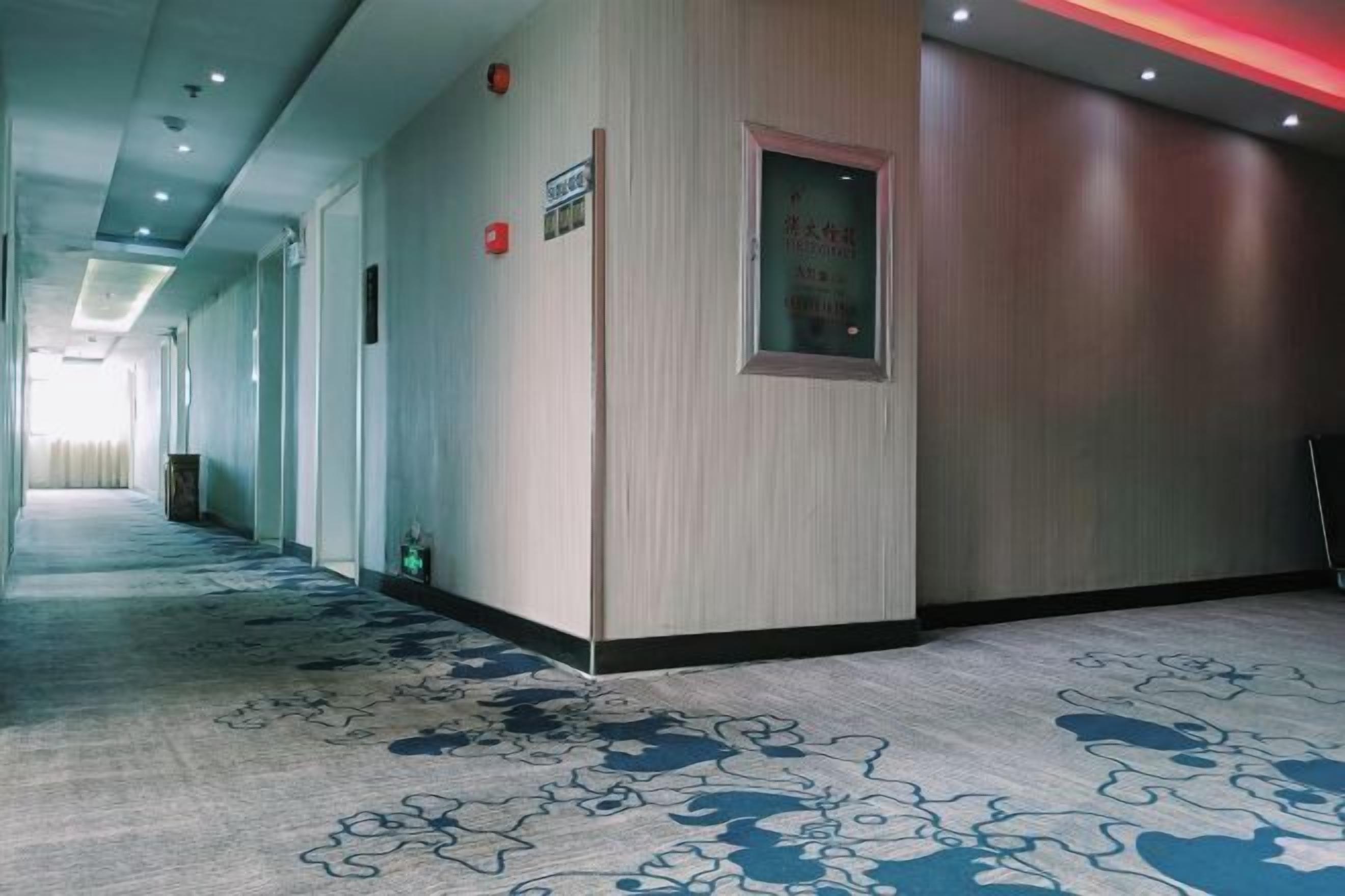 A hotel corridor with a patterned carpet, a wooden wall panel, and a door with a window.