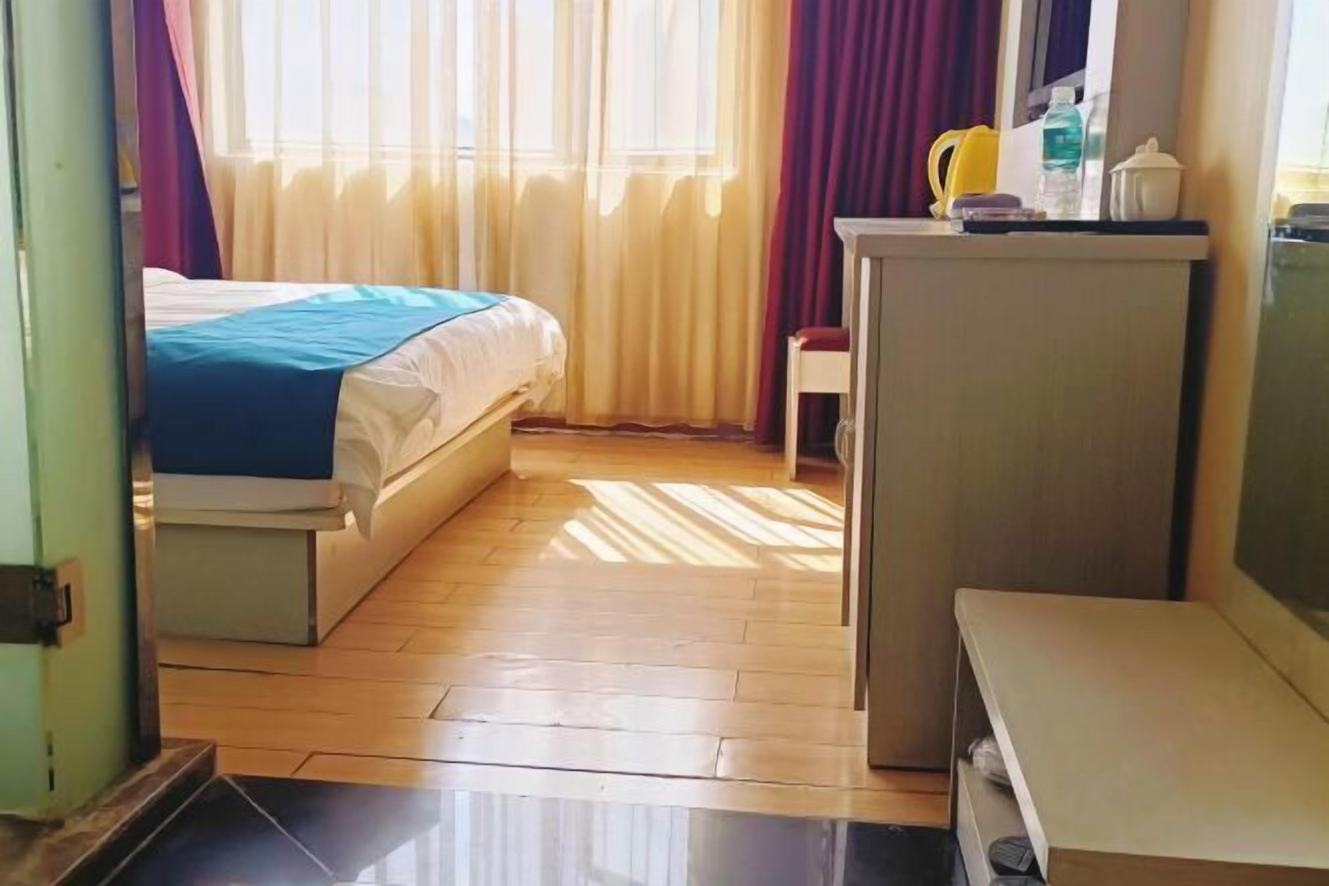 Quarto de hotel com uma cama, uma pequena escrivaninha, uma cadeira e uma janela com cortinas.