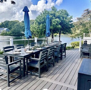 An outdoor dining area with a view of a lake, wooden deck, and blue umbrellas. - Sunset Point - Spacious 4-BR Long Pond House w/ private swimmable beach & AC (Yarmouth)