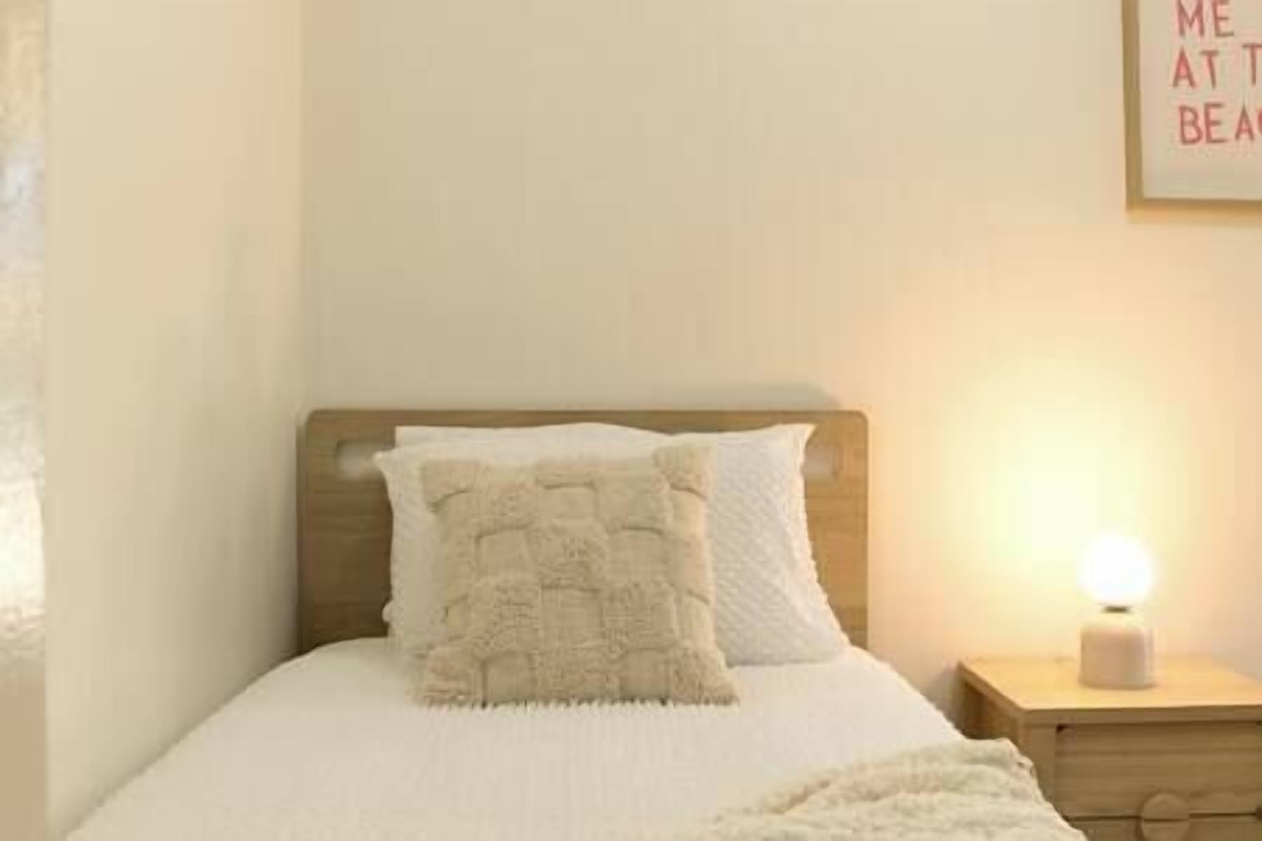 Un letto rifatto con copriletto bianco e cuscino decorativo beige, un comodino con una lampada, e una parete con un cartello parziale.