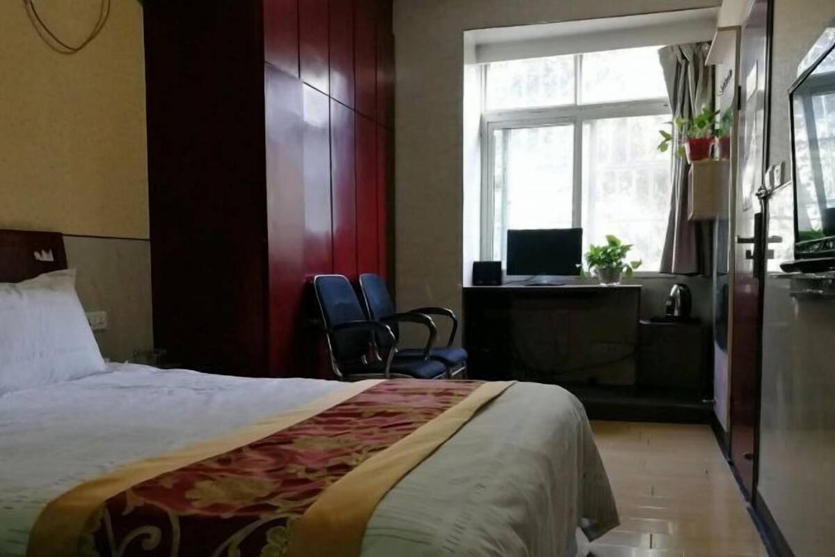 Habitación de hotel con cama, escritorio con computadora, dos sillas, ventana con cortinas y una planta pequeña.