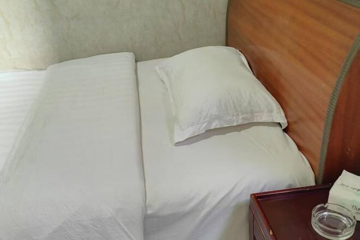 Una cama bien tendida con sábanas blancas, un cabecero de madera y una mesita de noche con un vaso y un papel.