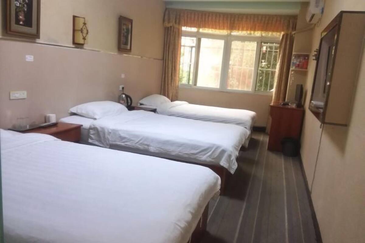 Habitación de hotel con tres camas individuales, una ventana con cortinas, una mesita con lámpara y un televisor montado en la pared.
