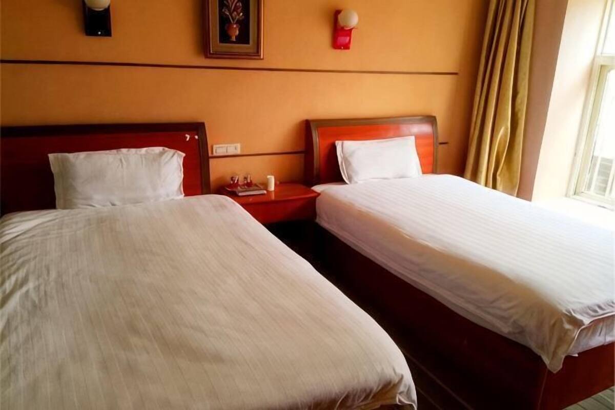 Dos camas individuales con cabeceros de madera, una mesita de noche con lámpara y una ventana con cortinas.