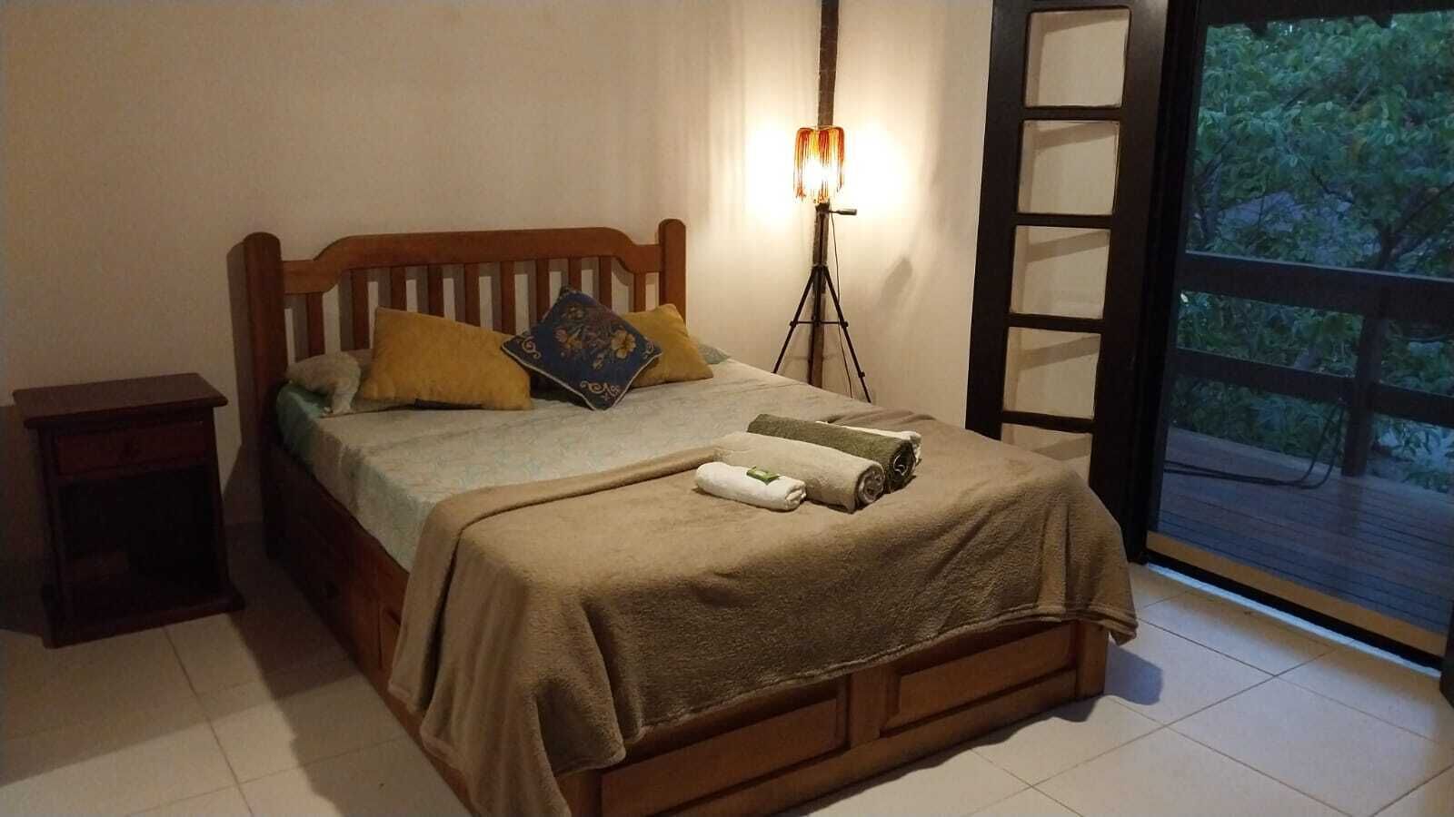 A bedroom with a wooden bed, a bedside table, a floor lamp, and a view of greenery through a window.