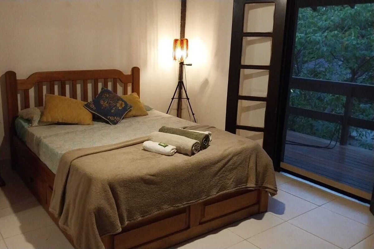 A bedroom with a wooden bed, a bedside table, a floor lamp, and a view of greenery through a window.