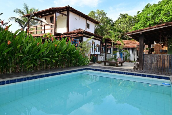 A swimming pool with tiled edges, surrounded by lush greenery and a building with a tiled roof. - Gambá Casa Boutique (Búzios)