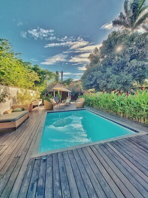 A rectangular pool with a wooden deck, surrounded by lush greenery and a thatched-roof gazebo. - Gambá Casa Boutique (Búzios)