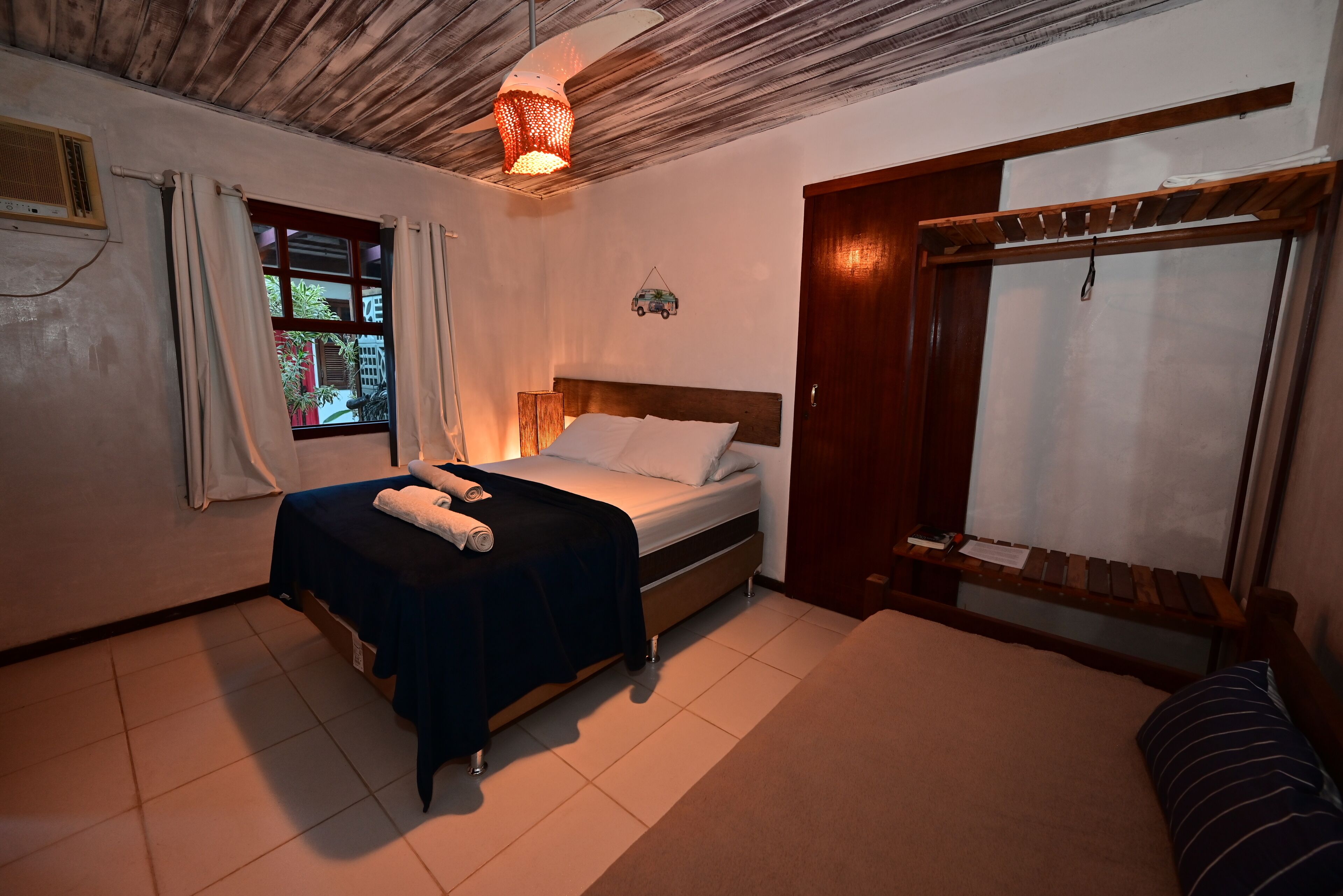 A bedroom with a bed, a window with curtains, a wooden ceiling, and a door with a wooden frame.