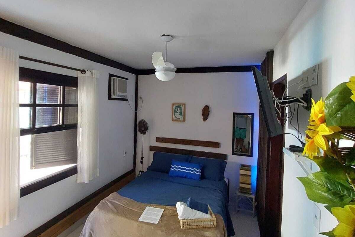 Ein Schlafzimmer mit einem Bett, einem Deckenventilator, einem Fenster mit Vorhängen, einem an der Wand montierten Fernseher und einer Pflanze.