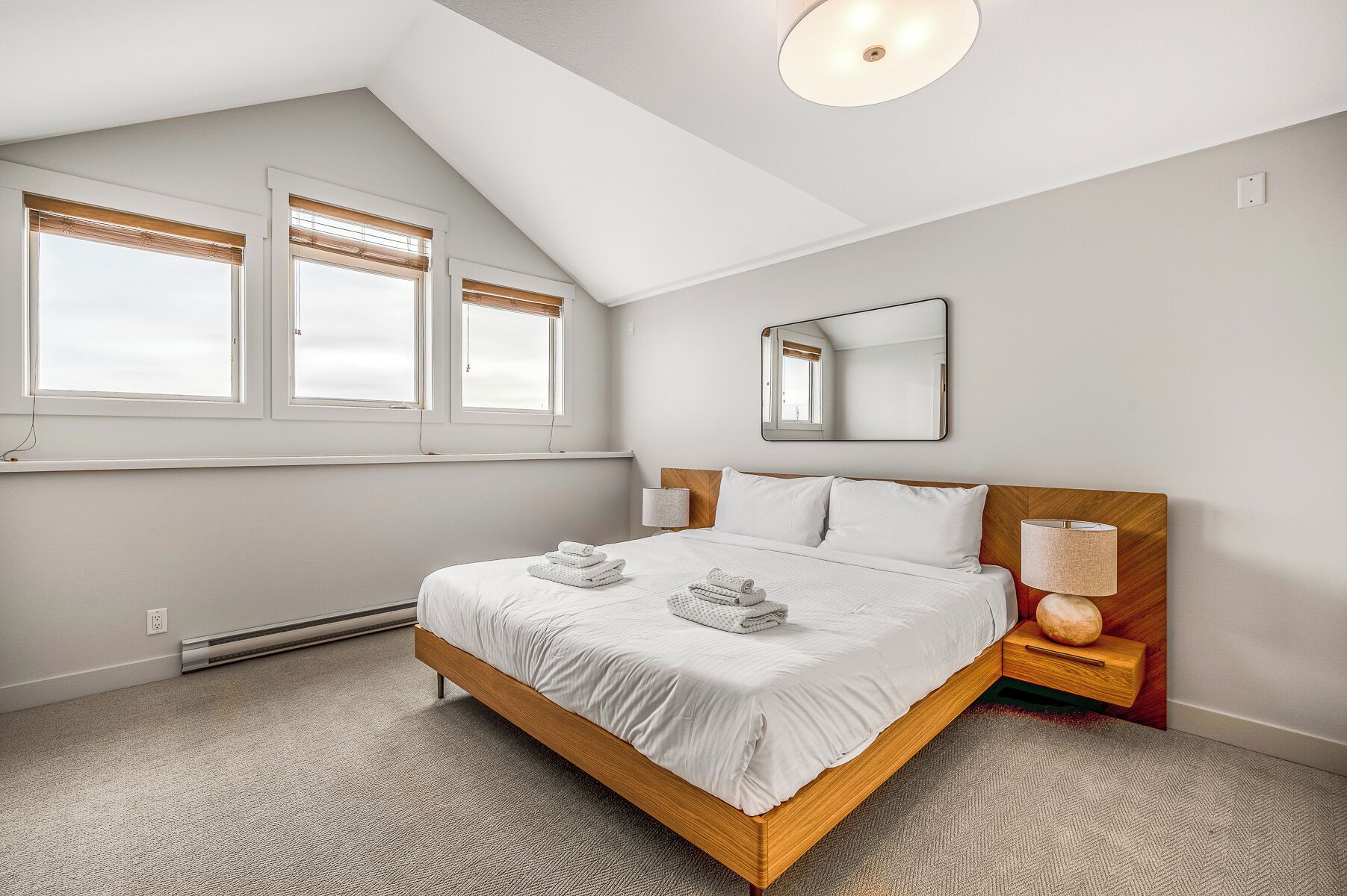 A modern bedroom with a sloped ceiling, a large bed with white linens, a wooden headboard, a bedside table with a lamp, and a window with blinds.