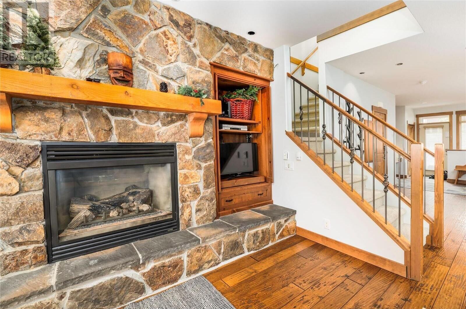 A stone fireplace with a TV shelf, a wooden staircase, and a wooden floor.