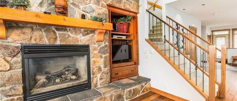 A stone fireplace with a TV shelf, a wooden staircase, and a wooden floor.