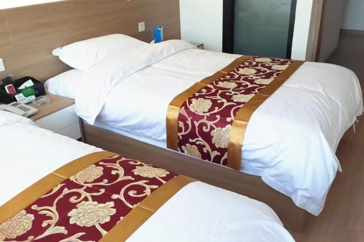 Une chambre d’hôtel avec deux lits, chacun agrémenté de draps blancs et d’une couvre-lit décorative rouge et or. On y trouve une table de chevet avec un téléphone et une bouteille d’eau.
