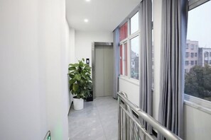 A narrow corridor with a potted plant, a door at the end, and a window with a view of buildings.