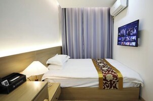 Habitación de hotel con cama, lámpara de noche, televisor montado en la pared y ventana con cortinas.