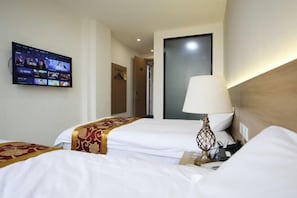 A hotel room with two beds, a TV mounted on the wall displaying a streaming service, a bedside lamp, and a door leading to another room.