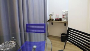 Une petite pièce avec une chaise bleue, un banc noir, une machine à café et une poubelle.