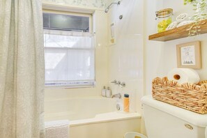 Ein Badezimmer mit Badewanne, einem Fenster mit Vorhang, einer Dusche und einem Ablagebereich mit Handtüchern und Pflegeartikeln.