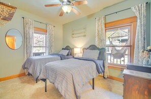 Ein Schlafzimmer mit zwei Betten, einem Deckenventilator, einem großen Fenster mit Vorhängen, einem Spiegel und einer Holzkommode.
