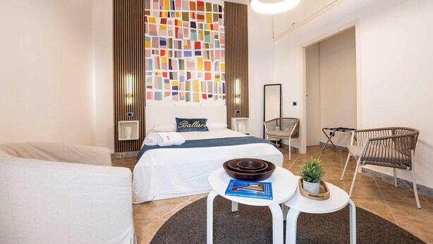 Een moderne hotelkamer met een bed, een klein tafeltje met een kom en een boek, een stoel, en een decoratief wandpaneel.