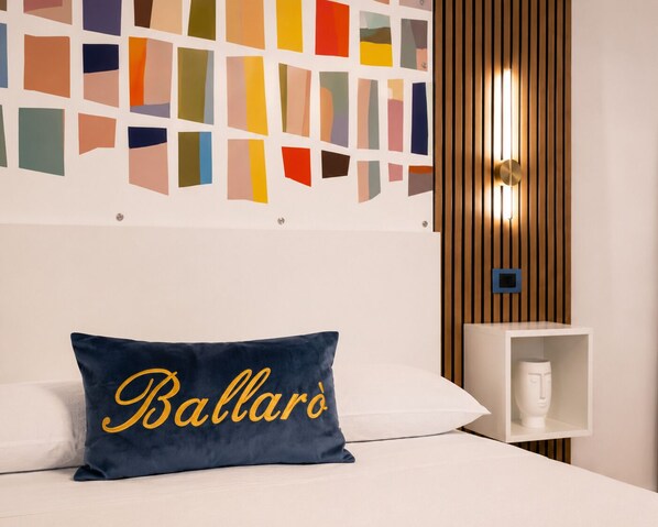 Une chambre d’hôtel avec un lit, un oreiller orné d’une broderie jaune représentant le mot « Ballarò », un mur décoré de motifs carrés colorés, un panneau mural en lattes de bois et une table de chevet blanche sur laquelle est posé un vase.