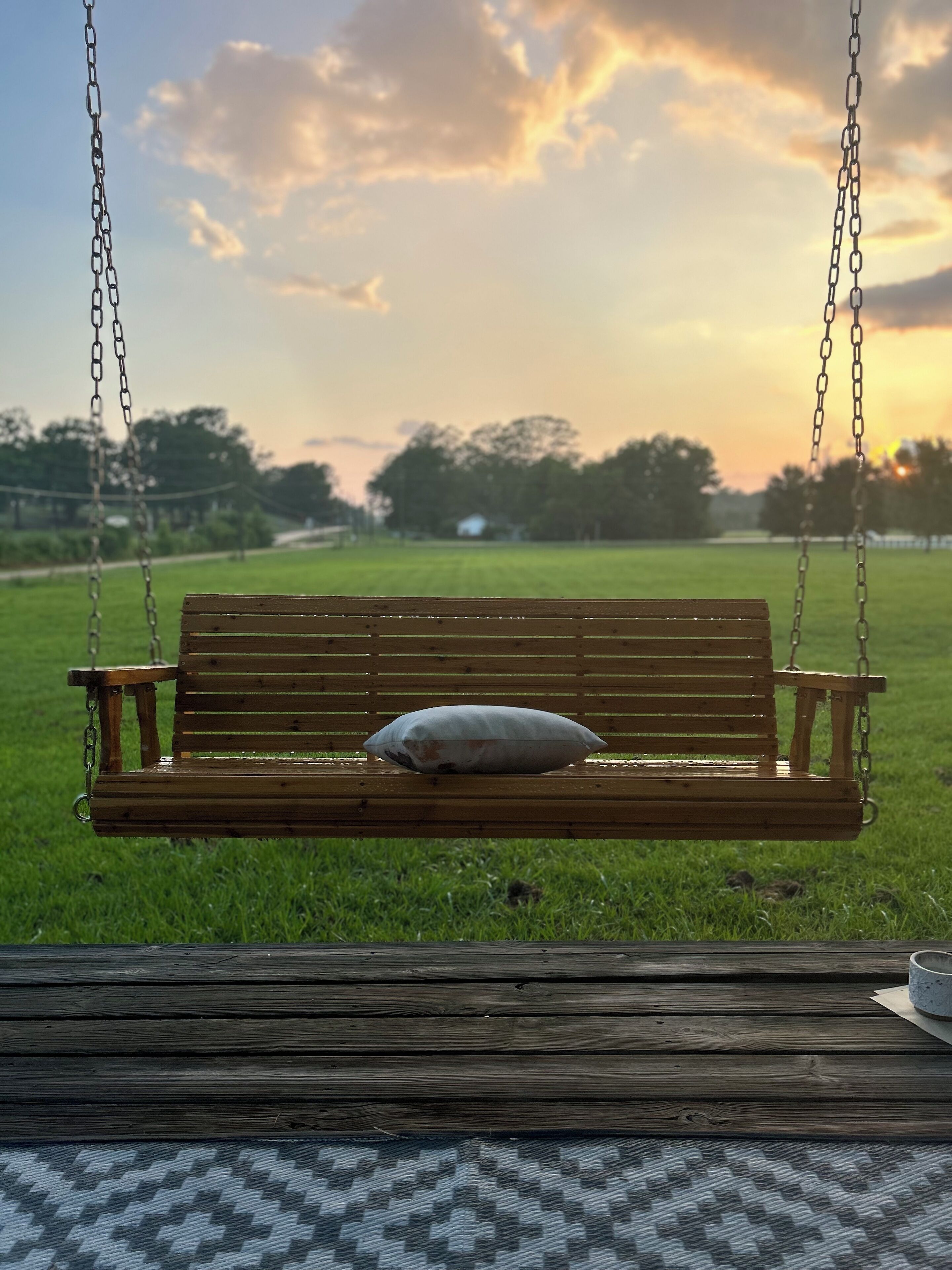 A wooden swing bench with a cushion, set in a grassy field with a sunset in the background.