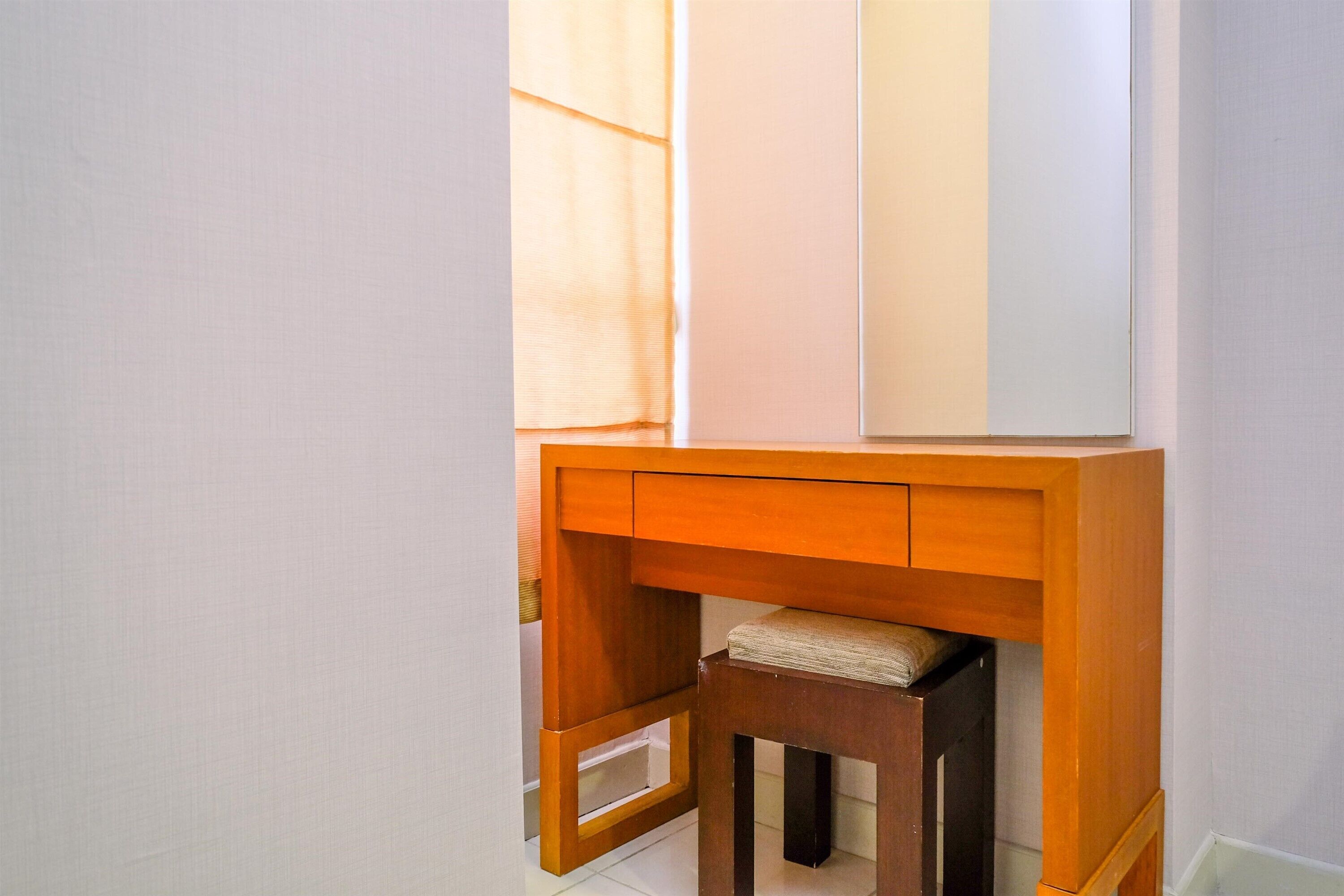 A corner of a room with a wooden desk and a chair, a window with blinds, and a white wall.