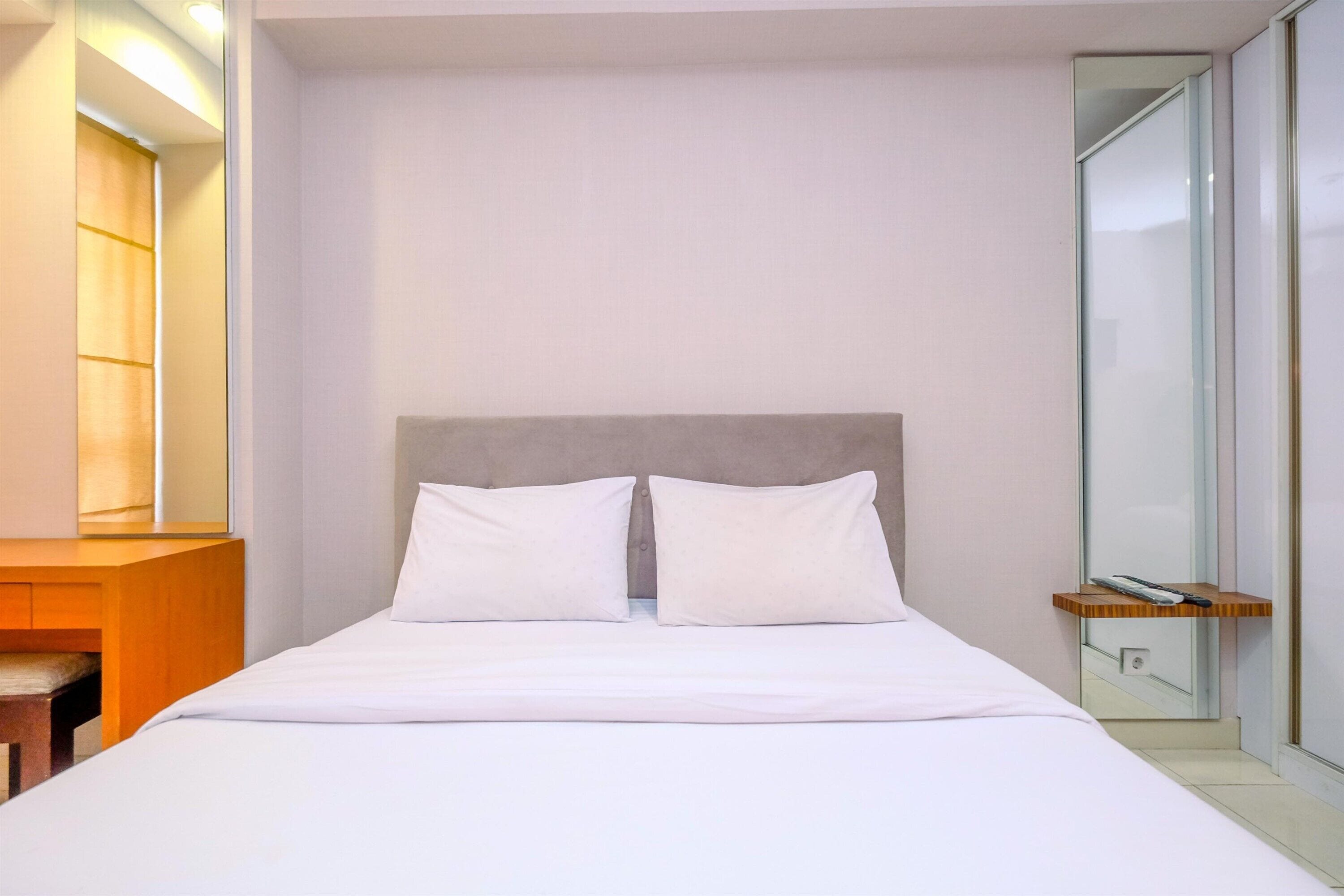 A modern hotel room with a large bed, a wooden desk, and a small shelf with a remote control.