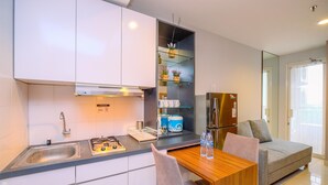 A modern kitchen with white cabinets, a stainless steel sink, and a built-in oven. There is a wooden dining table with chairs, a small refrigerator, and a glass shelf with plants. - Good And Comfy 2Br Cinere Bellevue Suites Apartment (Depok)