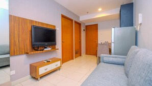 A modern living room with a flat-screen TV mounted on a wooden panel, a grey sofa, a wooden cabinet, and a refrigerator. - Good And Comfy 2Br Cinere Bellevue Suites Apartment (Depok)