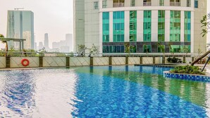 A swimming pool with a lifebuoy, surrounded by a low wall and plants. In the background, there are modern buildings with large windows. - Homey And Big Studio Apartment At The Wave Kuningan (Jakarta)