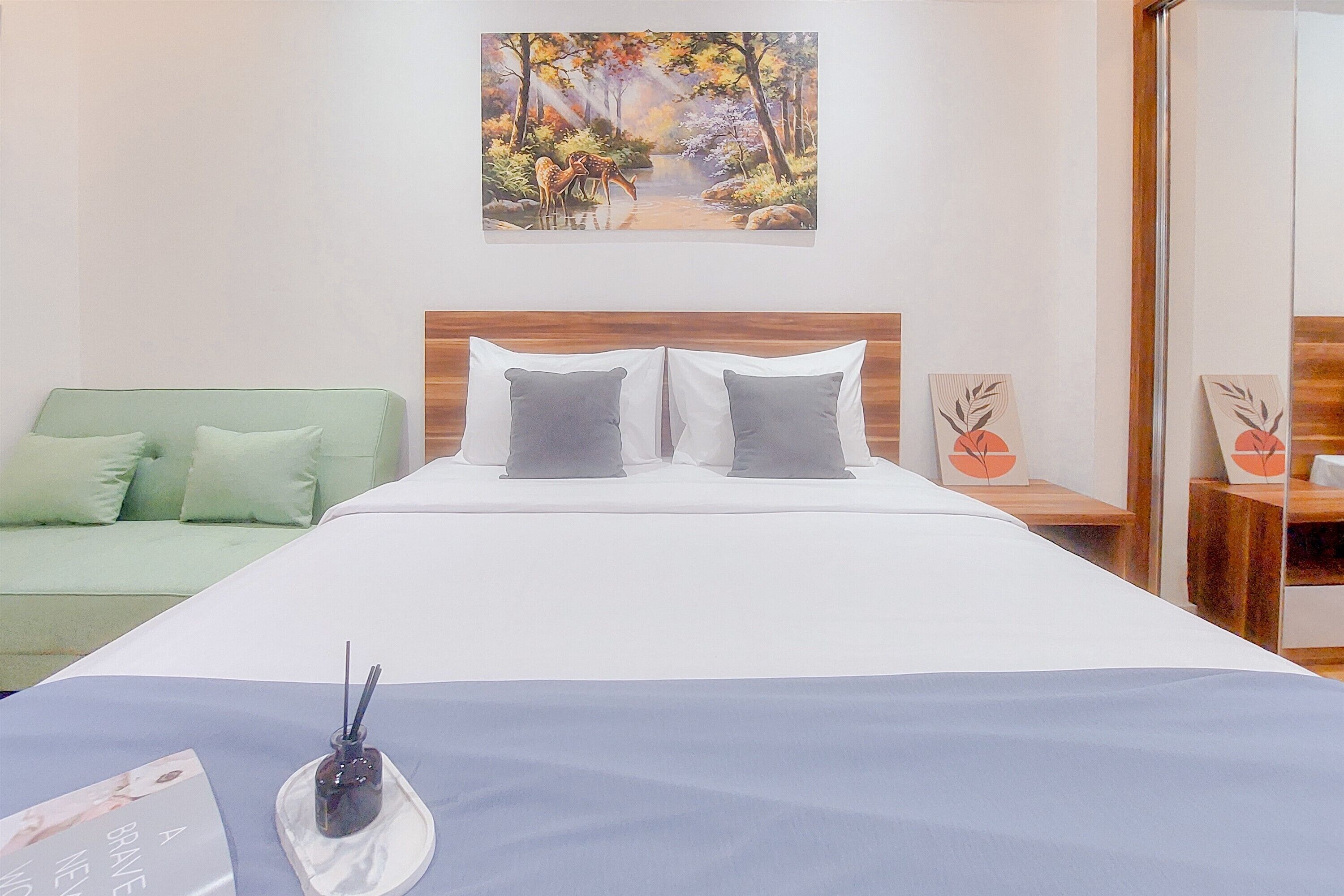 A hotel room with a bed, a green sofa, a painting of a forest scene, and a small table with a fruit design.