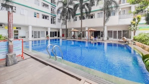A swimming pool surrounded by a multi-story building with balconies, palm trees, and outdoor seating areas. - Comfy And Homey Studio At Sky View Apartment (South Tangerang)