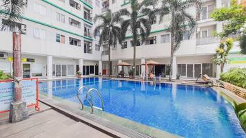 A swimming pool surrounded by a multi-story building with balconies, palm trees, and outdoor seating areas.